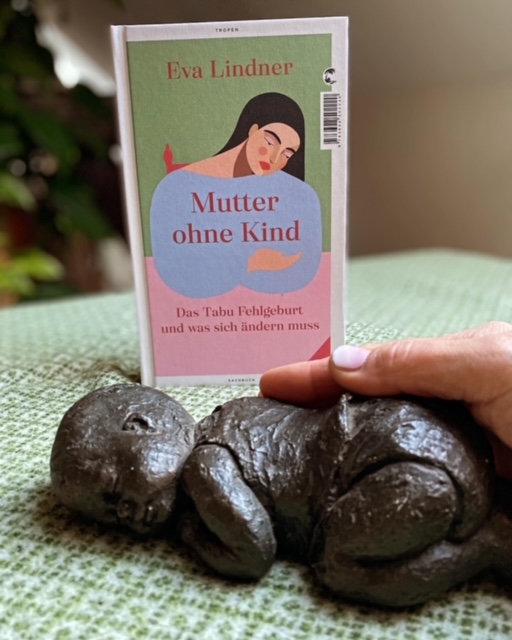 Ein Bild einer Statue mit einem Baby und einem Buch im Hintergrund: Titel: Mutter ohne Kind. Das Tabu Fehlgeburt und was sich ändern muss.