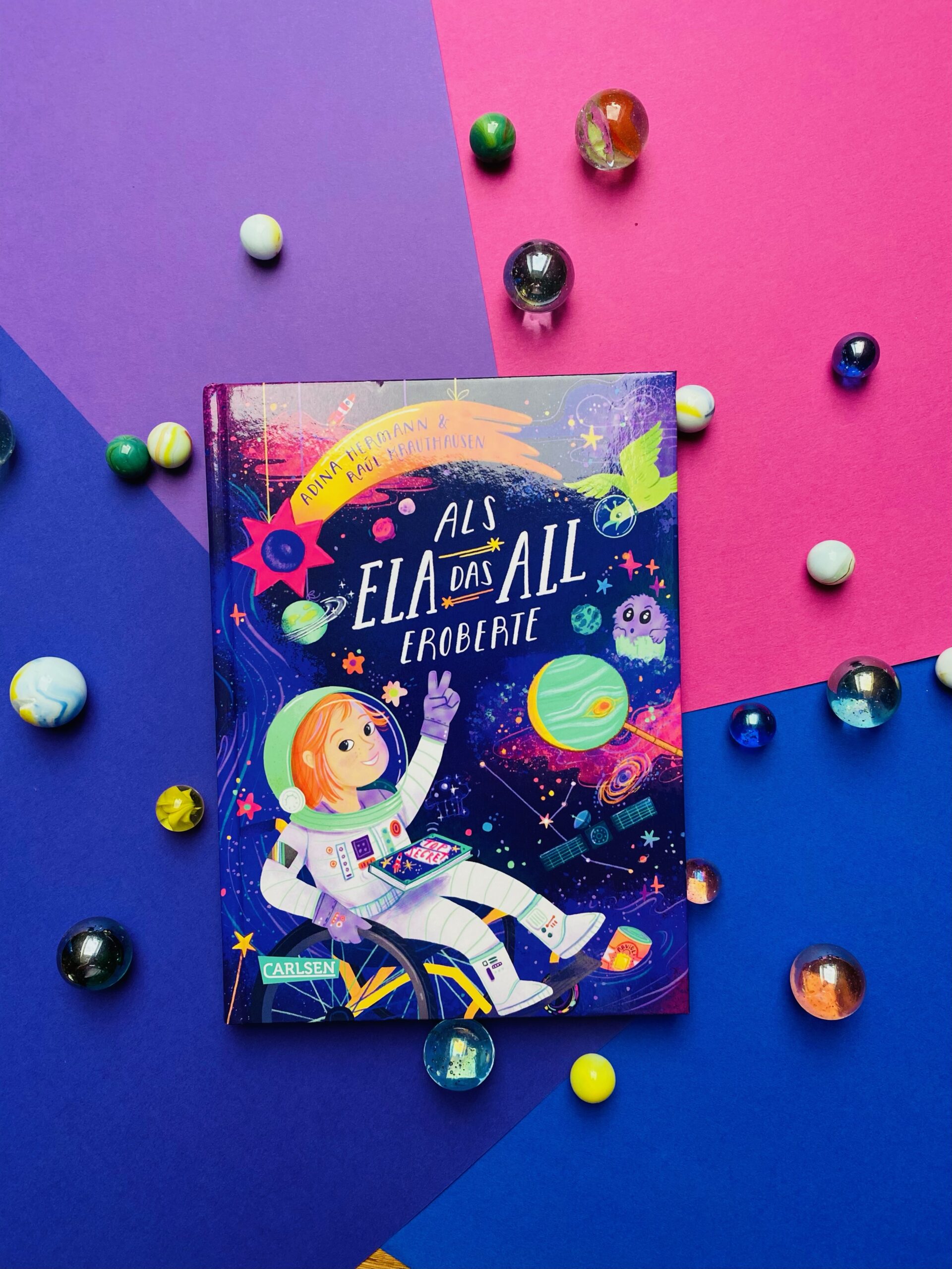 Bunter Hintergrund mit Murmeln und einem Buch: Als Ela das All eroberte. Ein farbenfrohes Buchcover, das Ela zeigt, die einen Astronautenanzug trägt und im All ist.